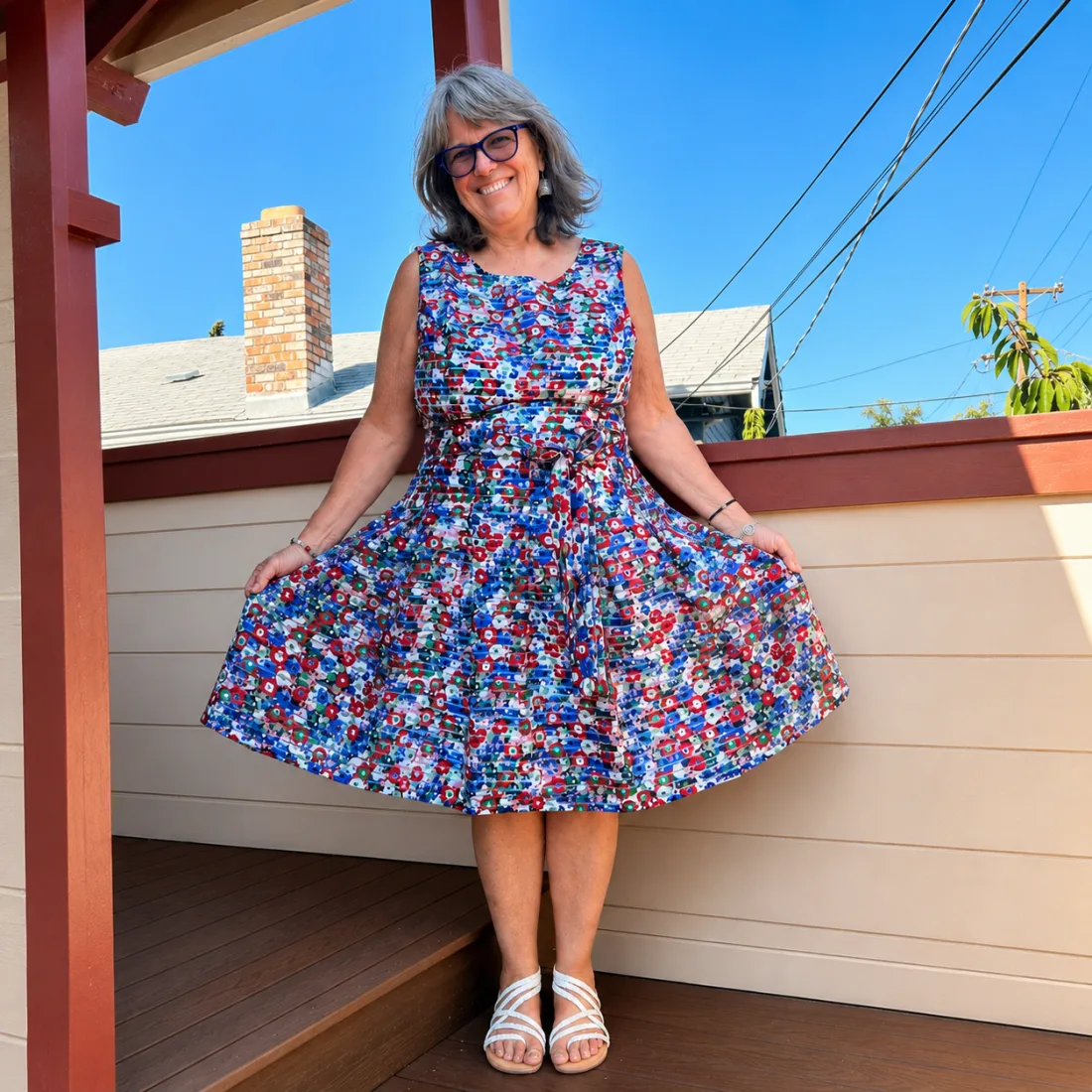 Anne &lsquo;Aunty Anie&rsquo; Symens-Bucher smiling on a sunlit porch at Canticle Farm in Oakland, holding out the skirt of a floral dress