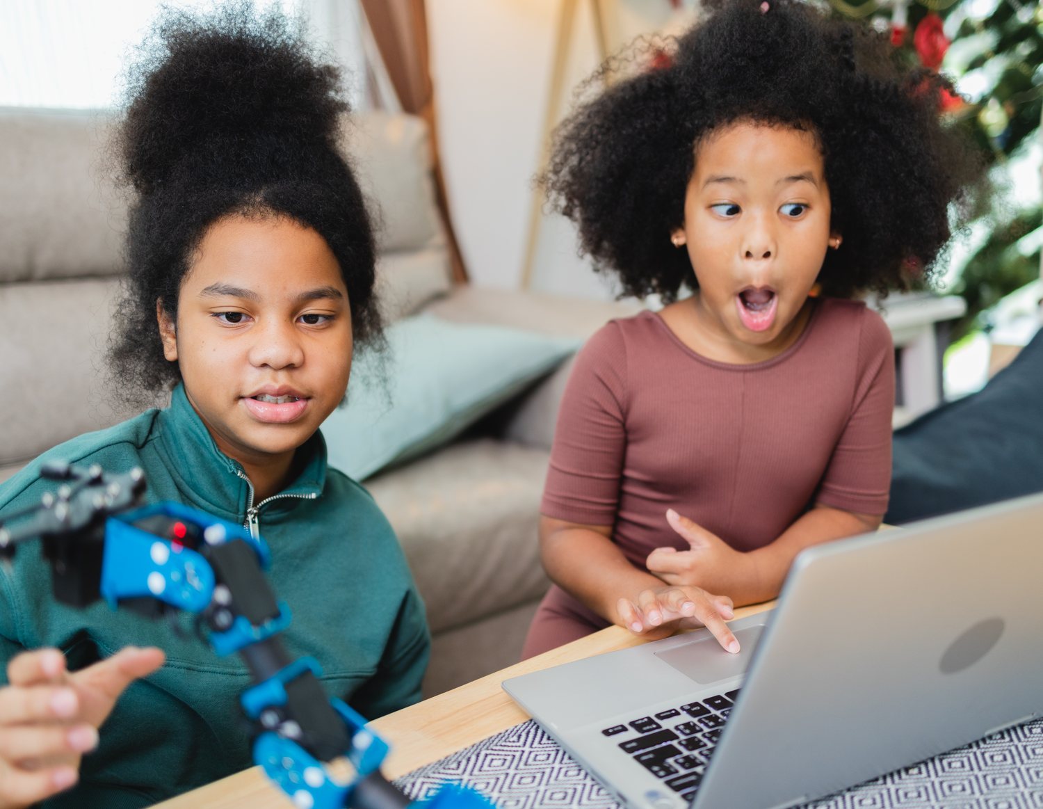 Two girls at a laptop with a robotic arm, one reacting with delight