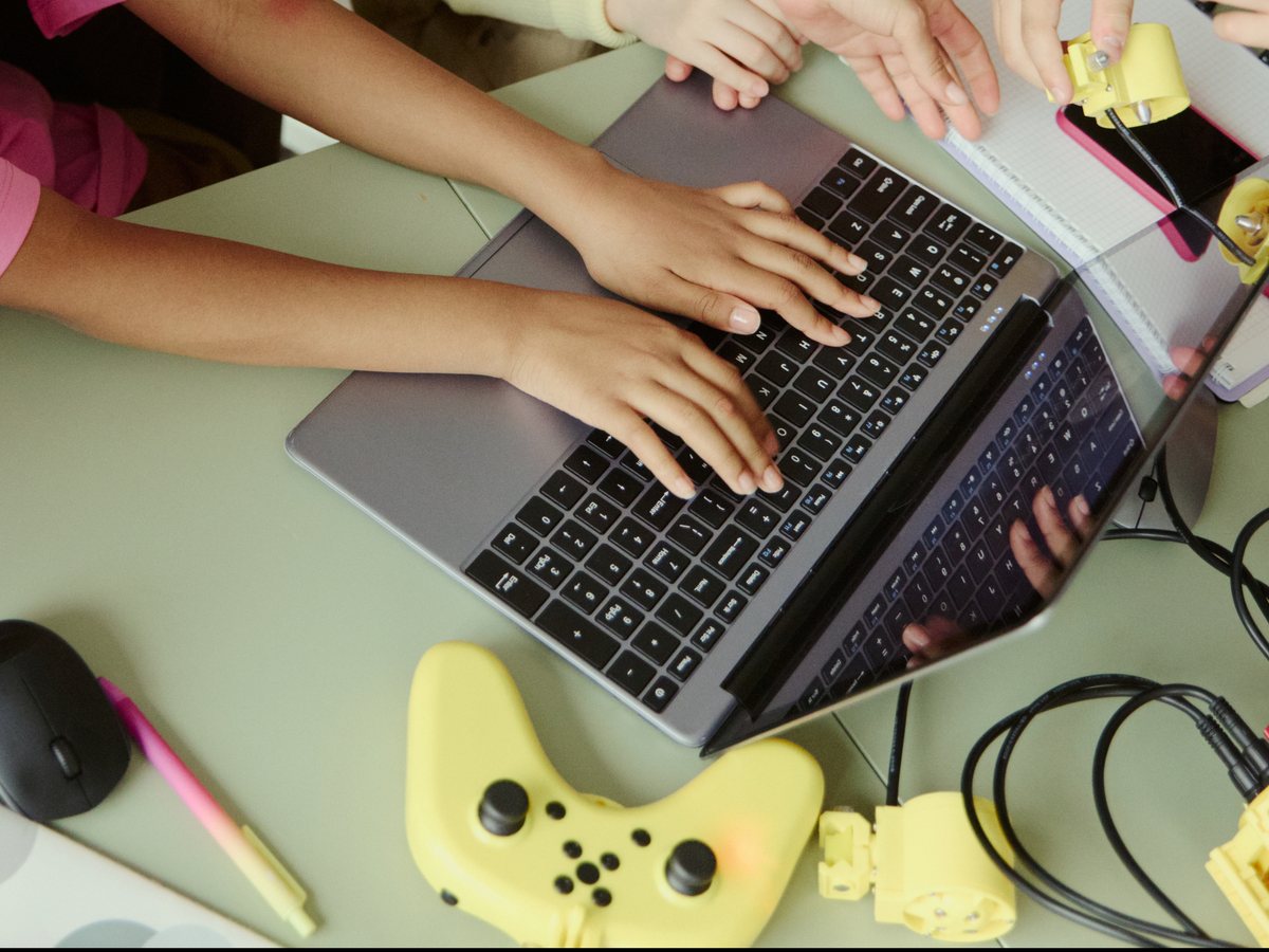 Children's hands coding on a laptop beside a handheld game controller